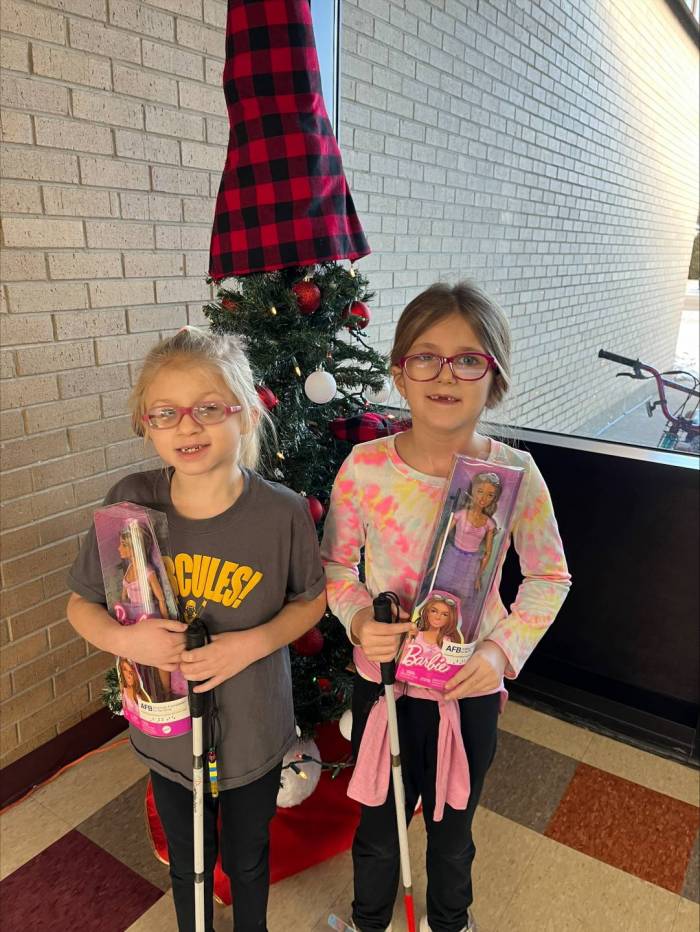 In front of a Christmas tree, two elementary aged girls proudly hold their own white canes as well as a box that holds a Barbie who also uses a white cane.